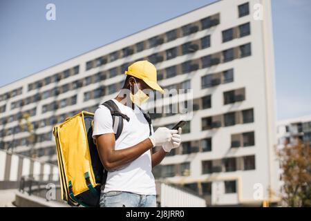 Attractive African American food delivery courier in face mask posing on the street. Smiling man in a yellow cap and with a heat-insulated food bag on his back with a phone in his hands. Stock Photo