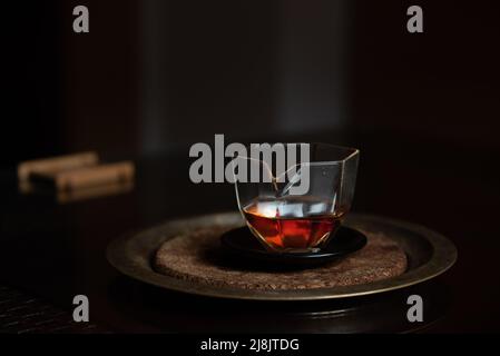 A small Chinese glass jug stands on a dark table against a dark background. There is caramel-colored tea in the pot. The pot is on a tray. Stock Photo