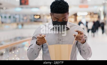 Close-up young african american man in protective medical mask standing in mall male client buyer birthday man looking into package with new purchase Stock Photo