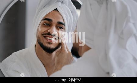 Funny millennial handsome man bearded indian arab guy with white bath towel on head looks into reflection in mirror bathroom good-looking male at home Stock Photo