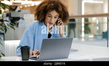 African tired exhausted businesswoman multitasking girl female worker freelancer talking on mobile phone finishing call looking at laptop holding head Stock Photo