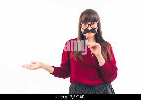 Dark-haired young woman holds fake moustache on a stick in front of her face and keeps her another hand open movember white background copy space isolated studio shot . High quality photo