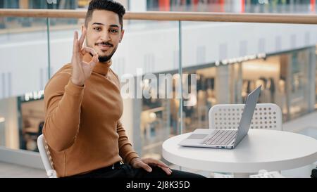 Young arab guy student freelancer typing on laptop surfing social network on internet admiring work new application showing approval sign consent Stock Photo