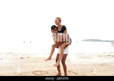 Asian man piggyback riding to a his couple in a sandy tropical beach Stock Photo