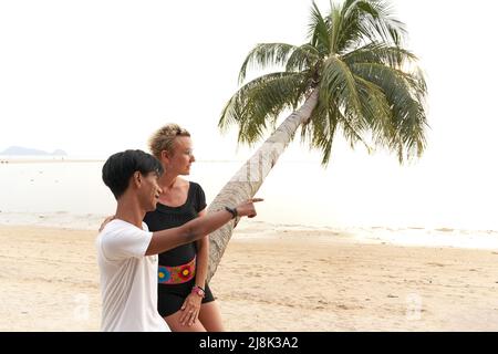 Young asian man pointing ahead next to a woman sitting on a palm tree Stock Photo