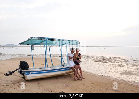 Couple sitting on a boat moored in the sand of a beach while drinking a coconut Stock Photo