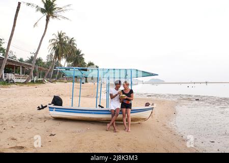 Lovers sitting on a boat moored in the sand of a beach while drinking a coconut Stock Photo