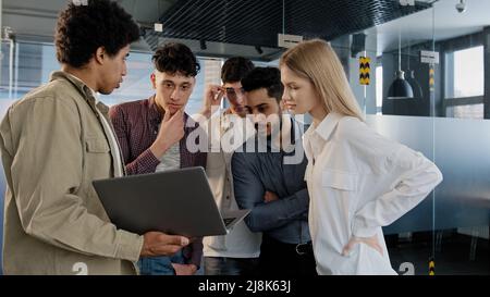 Young company employee explains development strategy tells new ideas showing work plan on laptop to smiling diverse colleagues creative team Stock Photo