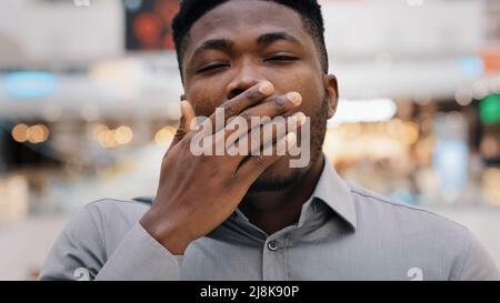 Male portrait close-up man overworked young african american man tired worker looking at camera yawns covers mouth with hand bored feels tired drowsy Stock Photo