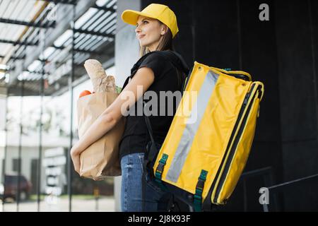 Delivery concept. Caucasian courier standing with a package with fresh food from the store. Smiling delivery woman holding paper bag with organic vegetables. Stock Photo