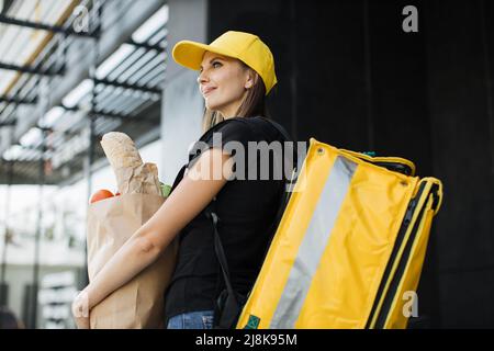 Delivery concept. Caucasian courier standing with a package with fresh food from the store. Smiling delivery woman holding paper bag with organic vegetables. Stock Photo