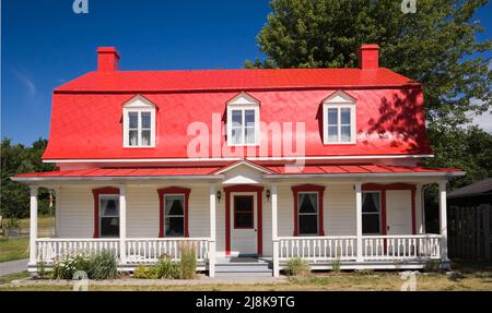 Old circa 1825 Canadiana cottage style home with white wood plank cladding and red sheet metal mansard roof in summer. Stock Photo