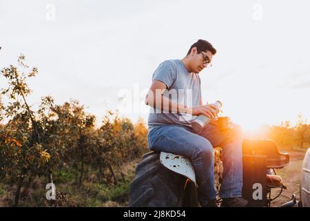 Young latin man drinking mate and relaxing, in a rural space, on top of an old little tractor. Hot beverage Stock Photo