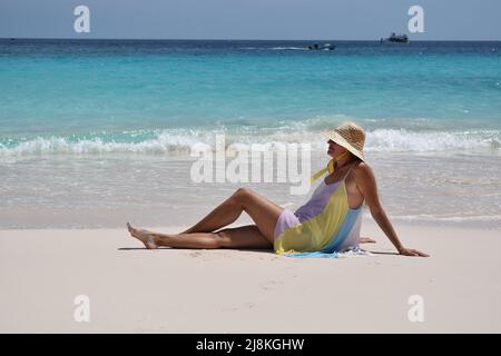 Lady wearing colourful light dress and straw hat on white sand beach with cruise ship on the horison. Stock Photo