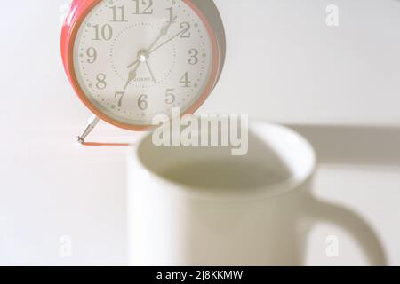 Red alarm clock with seven o'clock hands and a white cup with a hot morning drink. Selective focus on alarm clock Stock Photo