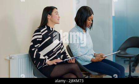 Asian and african american women talking in waiting lobby at job interview, preparing to join employment meeting with HR department. Female applicants having conversation about hiring. Stock Photo