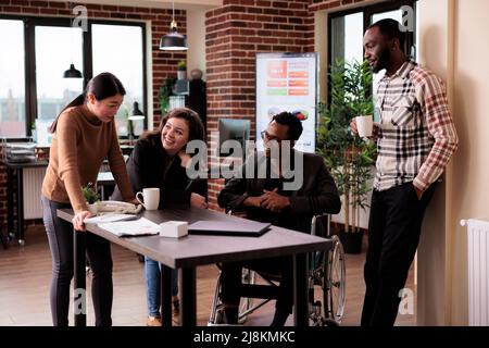 African american manager with physical disability laughing with coworkers, working in disability friendly office. Businesspeople and wheelchair user chatting on break in startup workplace. Stock Photo