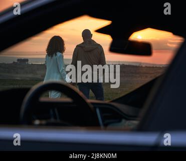 View through open side and front car window of couple in love. Back view of romantic pair looking at evening sky in orange and red colors while sunset out of city. Stock Photo