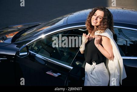 Happy woman standing by electric car on the street. Cheerful young lady with closed eyes wearing stylish white suit while posing near electric automobile outdoors. Stock Photo