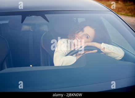 Beautiful woman looking at camera and smiling while sitting behind glass in electric automobile. Charming businesswoman resting head on steering wheel while sitting on driver seat in electro car. Stock Photo