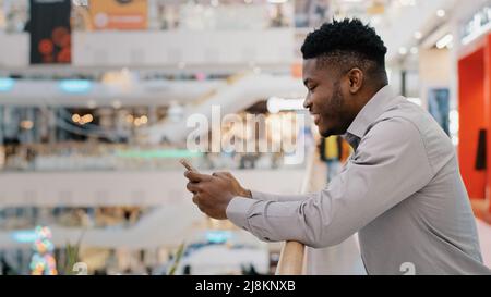 Young african american man standing in shopping mall spending free time on social media internet on smartphone using virtual service pleasant Stock Photo