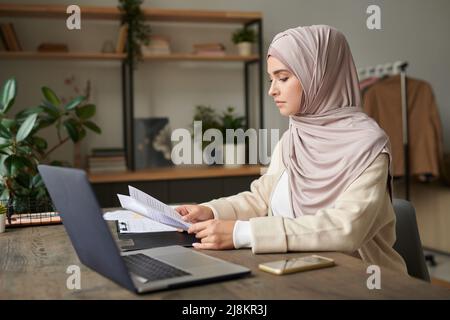 Serious Muslim woman wearing hijab sitting at desk in modern office looking through important documents Stock Photo
