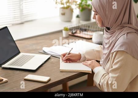 Successful Muslim woman wearing headscarf making notes in her copybook while working on business plan Stock Photo