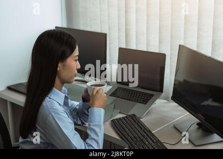 Asian woman was drinking coffee in the early morning while contemplating the program and code on the computer screen. Stock Photo