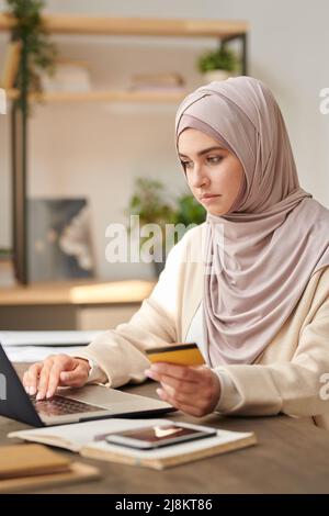 Vertical medium portrait of modern young Middle Eastern woman wearing hijab holding credit card buying goods in online shop Stock Photo