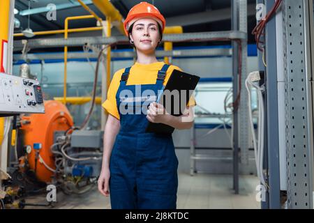 A female worker in helmet and uniform with a folder in her hand. In the background, boiler equipment and pipes. Concept of industrial production and f Stock Photo