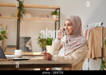 Modern Muslim woman working as fashion designer sitting at desk in cozy office room talking on phone Stock Photo
