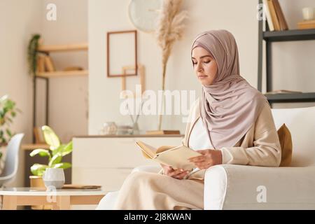Beautiful Muslim woman wearing pale pink hijab and casual clothing sitting relaxed in armchair reading book, copy space Stock Photo