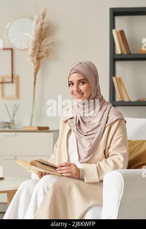 Vertical portrait of joyful young Middle Eastern woman wearing hijab sitting in armchair holding book smiling at camera Stock Photo
