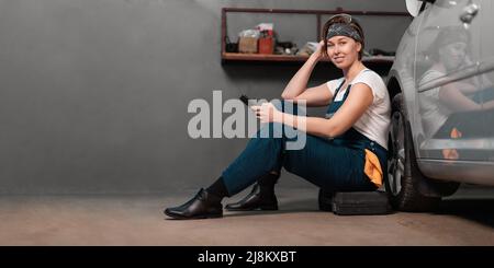 Banner of women's equality. Smiling caucasian young woman mechanic is sitting leaning on a car in an auto repair shop and using cellphone. Copy space. Stock Photo