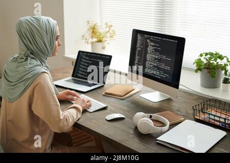 Horizontal high angle shot of modern Muslim woman wearing hijab sitting at table working on computer code, copy space Stock Photo