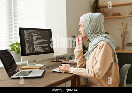 Successful Muslim programmer wearing hijab sitting at desk working on new software using desktop PC and laptop, copy space Stock Photo