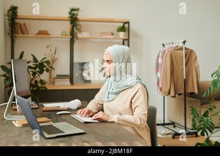 Serious young Muslim professional programmer wearing light blue hijab sitting at desk at home working on software Stock Photo