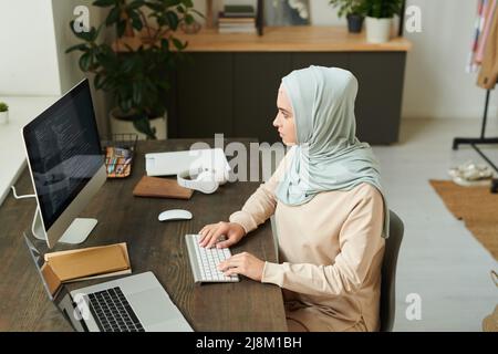 Horizontal high angle view shot of modern young Muslim woman working on programming using desktop computer Stock Photo