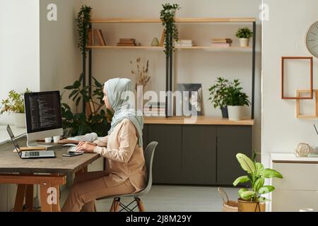 Young Middle Eastern professional programmer wearing light blue hijab sitting at desk at home working on new computer software, copy space Stock Photo