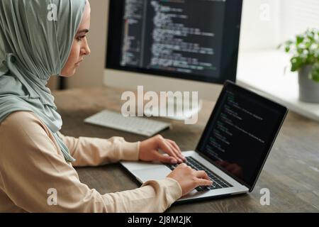 Professional programmer wearing hijab sitting at office table typing computer code on laptop, copy space Stock Photo