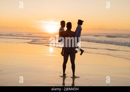 Rear view of african american young man picking up son and daughter on beach against sky at sunset Stock Photo
