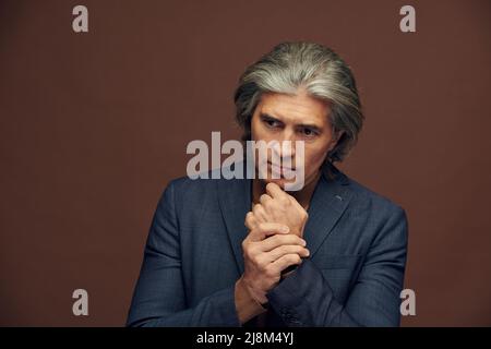 Portrait of serious curious mature businessman in stylish jacket touching chin while posing against isolated background Stock Photo