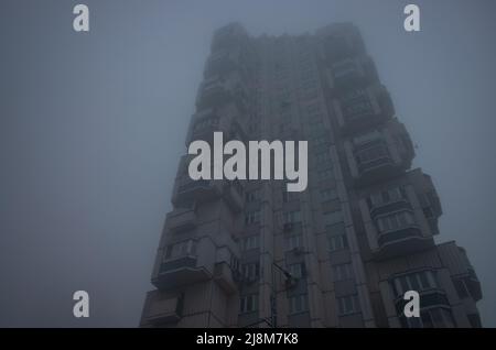 Tall residential building in the form of a tower, immersed in a misty gray sky. Cyberpunk stylistics. Dramatic, depressing mood. Stock Photo