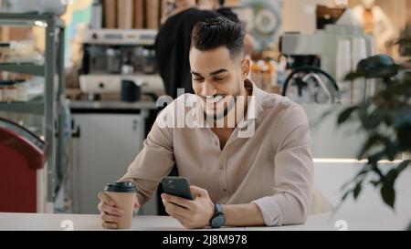 Indian hispanic arabian man multiracial businessman sitting at cafe table drinking hot coffee tea from paper cup looking in mobile phone typing Stock Photo