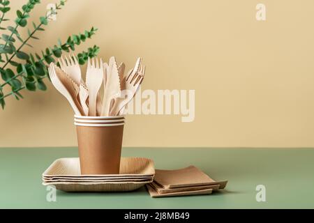 Disposable dishes and utensils against beige green background. Biodegradable wooden cutlery in brown paper cups on plates. Eco-friendly tableware. Stock Photo