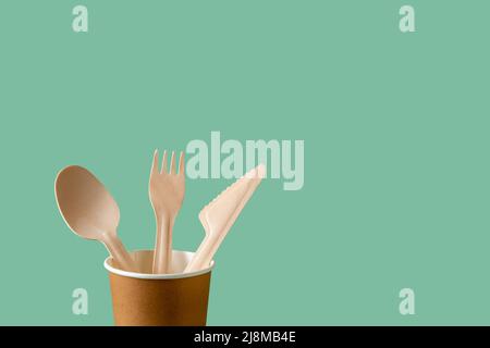 Disposable wooden spoon, fork and knife in a brown paper cup against green background. Biodegradable tableware for takeaways, picnics, fast food. Stock Photo