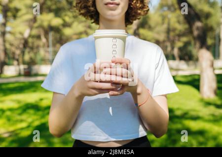 Young redhead woman holds paper cup of hot coffee, closes eyes, wears white tee, green city park background, outdoors. Summer fun, enjoying life, vaca