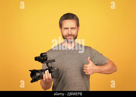 cheerful bearded man videographer making movie for vlog with camcorder showing thumb up, filming Stock Photo