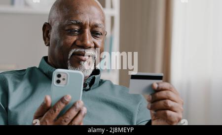 Elderly African Dominican old mature man at home online transaction buying hold smartphone credit card successful payment buy on internet with mobile Stock Photo