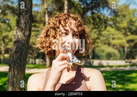 Happy redhead woman eating protein bar at city park, outdoors and looking away. Nutrition for sport. Protein bar as she takes a break from her daily j Stock Photo